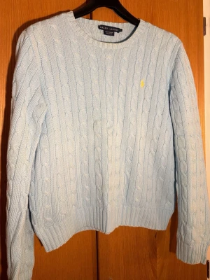 Ralph Lauren Ljusblå kabelstickad tröja Ralph Lauren - Supersnygg ljusblå kabelstickad tröja från Ralph Lauren med gul broderad logga på bröstet. Klassisk rund halsringning och långa ärmar. Perfekt till både jeans och kjol för en clean look.