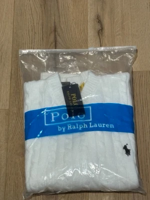 Vit kabelstickad tröja Polo Ralph Lauren - Vit kabelstickad tröja från Polo Ralph Lauren med klassisk rund hals och svart broderad logga på bröstet. Tröjan har långa ärmar och ribbade muddar vid ärmslut och nederkant. Perfekt för dig som gillar stilren och tidlös design.