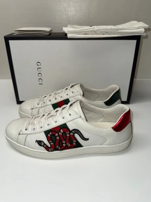 Gucci Ace sneakers - Gucci Ace sneakers i vitt skinn med ikoniska gröna och röda ränder på sidan och broderad orm. Baksidan har metallicröd och metallicgrön detalj i ormskinnslook. Klassisk låg modell med snörning och rund tå. Snygg statement-sko för dig som gillar lyxiga detaljer.