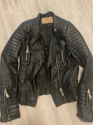 Svart skinnjacka från Vojelles, strl 36 - Säljer en svart skinnjacka från Vojelles i storlek 36. Jackan har coola detaljer som dragkedjor, bälte i midjan och vadderade partier på axlar och ärmar. Snygg bikerstil som passar perfekt till jeans eller kjol.