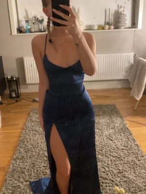 Marinblå balklänning med snörning - Säljer en oanvänd marinblå balklänning med tunna axelband och djup slits framtill. Klänningen har öppen rygg med snygg snörning som korsas och knyts baktill. Materialet är glansigt och följsamt, perfekt för bal eller fest.