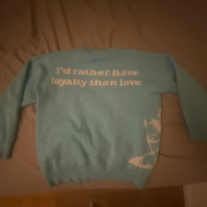 Blå stickad tröja med vit text - Säljer en ljusblå stickad tröja med vit text på ryggen: 'I'd rather have loyalty than love'. Framsidan har en cool illustration av en amor med pilbåge och ett brustet hjärta. Rund hals och långa ärmar. Jag är 175cm och den är lite liten.