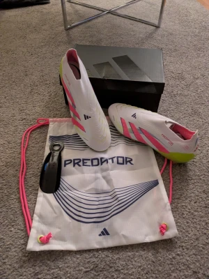 Adidas Predator Elite LL F fotbollsskor - Säljer ett par Adidas Predator Elite LL F fotbollsskor i storlek 46. Skorna är vita med rosa och limegula detaljer, har en strömlinjeformad design utan snörning och en sula med dobbar för gräsplan. Tillbehör som påse och skohorn ingår. Helt nya, ny pris typ 2000krb
