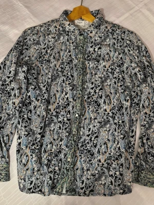 Mönstrad skjorta från Saint Tropez XS - Unik långärmad skjorta från Saint Tropez i storlek XS. Skjortan har ett detaljerat paisleymönster i svart, vitt, blått och gult samt snygga tryckknappar framtill och vid ärmsluten. Tillverkad i mjuk bomull som känns skön mot huden.