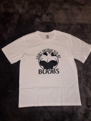 ”Easily distracted by boobs” T-shirt  - Säljer en vit t-shirt med svart tryck och texten 'Easily distracted by boobs' på bröstet. Klassisk passform med rund hals och korta ärmar. Perfekt för dig som gillar statement-plagg och vill sticka ut lite extra.