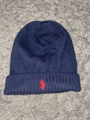 Mörkblå stickad mössa från Polo Ralph Lauren - Mörkblå stickad mössa från Polo Ralph Lauren med klassisk röd broderad logga framtill. Mössan har uppvikt kant och är gjord i ett mjukt och värmande material som passar perfekt för kyliga dagar.Storlek: One size. Ca 18 cm bred platt och ca 23 cm hög. Stretchig modell som passar de flesta.