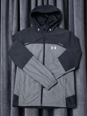 Under armour jacka  - Snygg tvåfärgad vindjacka från Under Armour i mörkgrått och ljusgrått. Jackan har huva, dragkedja framtill och logga på bröstet. Perfekt för blåsiga dagar och har en sportig look med långa ärmar och praktiska detaljer.