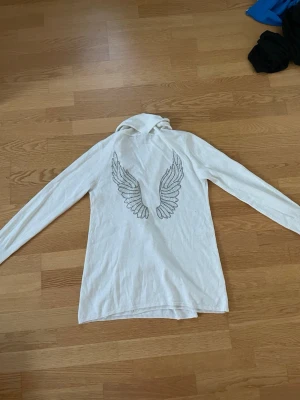 Vit kofta med vingar på ryggen från AVA - Säljer en vit långärmad kofta från AVA by Paul Stragas. Vingar på ryggen av rhinestones.