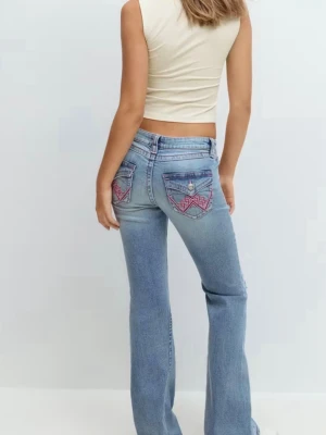 Bootcut ljusblå jeans med broderi - Säljer ett par* ljusblå bootcut jeans med slitningar framtill och rosa broder - crossing på bakfickorna. Lågmidjade och med klassisk femficksdesign. Jeansen har en avslappnad passform och är tillverkade i jeansmaterial.