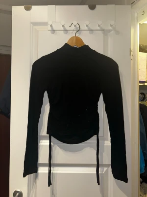 Svart långärmad polotopp från BikBok - Svart långärmad polotopp från BikBok i ribbat material. Toppen har snörning i sidorna som ger en snygg, justerbar passform och en croppad look. Perfekt att styla med jeans eller kjol för en trendig outfit.