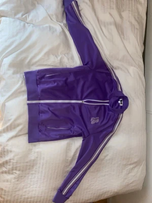 Lila track jacket från Risky - Säljer en lila track jacket från Risky i storlek M. Jackan har vita ränder längs ärmarna, dragkedja framtill och broderad R-logga på bröstet. Tillverkad i glansig polyester med ribbade muddar och fickor med dragkedja. Perfekt för en sportig och trendig look.
