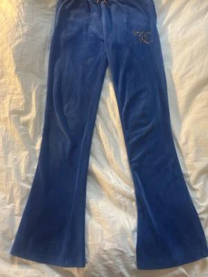Blå Juicy Couture byxor - Säljer ett par fina Juicy Couture byxor  med bootcut-modell och snörning i midjan med gulddetaljer.Byxan har ett glittrigt JC-logotypmärke på höften men dessvärre har några diamanter trillat av. byxorna  är storlek 10-11 år men funkar som xxs. Äkta🩷