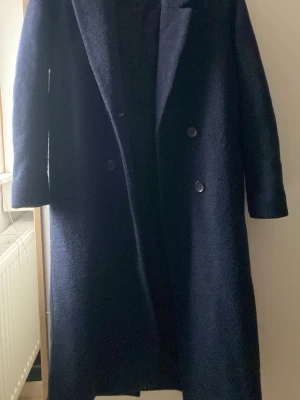 Mörkblå ullkappa från Monki XS - Lång, mörkblå kappa från Monki i storlek XS. Klassisk dubbelknäppt design med stora slag och raka ärmar. Tillverkad i ullblandning som håller dig varm under kyliga dagar. Perfekt oversized look för dig som gillar stilrena och tidlösa plagg.