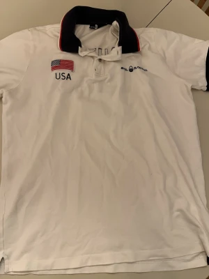 Vit Sail Racing USA piké - Säljer en vit pikétröja från Sail Racing med broderad USA-flagga och texten 'USA' fram samt 'UNITED STATES' och flagga bak. Svart krage med röd och vit kant, samt broderad Sail Racing-logga på bröstet. Klassisk och sportig look.