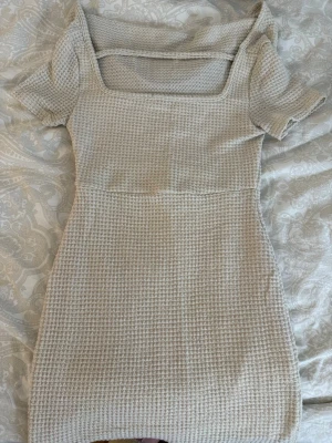 Beige ribbad kortklänning - Säljer en beige kortklänning med ribbad struktur och fyrkantig urringning. Klänningen har korta ärmar och är figurnära i modellen. Perfekt för dig som gillar en enkel men snygg stil. Materialet känns mjukt och stretchigt.