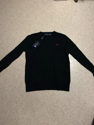 Svart kabelstickad tröja Polo Ralph Lauren - Svart kabelstickad tröja från Polo Ralph Lauren med klassisk rund hals och röd broderad logga på bröstet. Tröjan har lång ärm och ribbade muddar vid hals, ärmslut och nederkant. Perfekt för dig som gillar stilrena och tidlösa plagg. (Lite lite i storlek) passar M) 