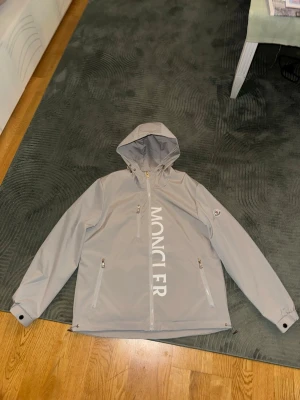 Grå vindjacka från Moncler - Snygg grå vindjacka från Moncler med stor logga framtill och patch på ärmen. Jackan har huva, tre dragkedjeförsedda fickor och justerbara muddar. Materialet är lätt och känns lyxigt, perfekt för dig som vill ha en clean och modern look.