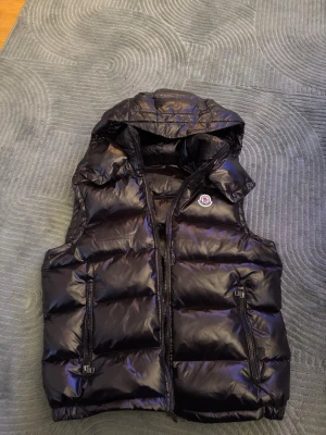 Svart dunväst från Moncler  - Säljer en svart dunväst från Moncler med glansig finish och avtagbar huva. Västen har två fickor med dragkedja, quiltad design och klassisk Moncler-logga på bröstet. Perfekt för lager-på-lager och riktigt snygg till streetwear-stilen.              Storlek L men passar perfekt till storlek M