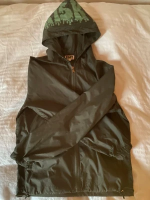 Grön vindjacka med huva från Risky - Säljer en mörkgrön vindjacka från Risky i storlek S. Jackan har huva med tryck, dragkedja framtill och en cool grön detalj på ärmen. Perfekt för dig som gillar streetwear och vill ha något lätt och snyggt till vardags. Snören till jackan saknas.