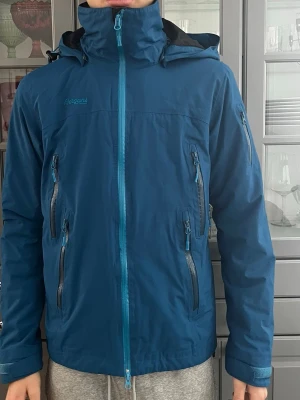 Blå Skidjacka från Bergans of Norway. Stl S - Säljer en blå skidjacka från Bergans of Norway. Jackan är fodrad med justerbar huva, snölås och flera dragkedjeförsedda fickor. Ärmsluten har justerbara ärmslut och snygga detaljer som broderad logga på ärmen. Jackan är i nyskick. Kragen är lite nopprig på insidan. Perfekt för skidåkning eller på fritiden. Personen på bilden är 186cm lång..