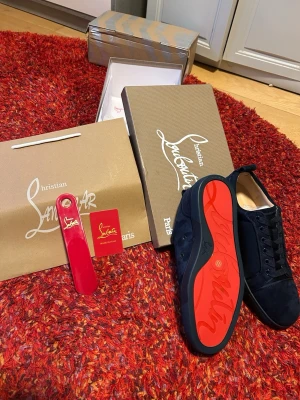 Mörkblå sneakers från Christian Louboutin - Säljer ett par mörkblå sneakers från Christian Louboutin i mocka med den ikoniska röda sulan. Skorna har rund tå, snörning och diskret broderad logga på hälen. Kommer med originalkartong, påse och tillbehör. Perfekta för dig som vill sticka ut med stil.
