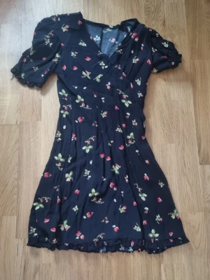 Svart blommig omlottklänning - Supersöt svart kortklänning med blommigt mönster och små jordgubbar. Klänningen har puffiga kortärmar, v-ringning och omlottdetalj framtill. Volangkant nertill ger en extra gullig touch.