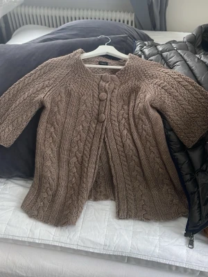 Brun stickad kofta från Lindex - Mysig brun stickad kofta från Lindex i storlek S. Koftan har flätstickat mönster, stora dekorativa knappar framtill och trekvartsärm. Perfekt för lager på lager under höst och vinter. Snygg att slänga över en t-shirt eller topp.
