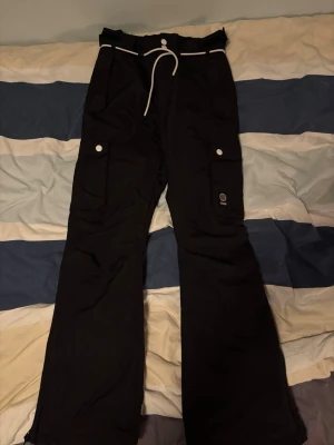 Svarta täckbyxor från Dope - Svarta täckbyxor från Dope med vita detaljer och snörning i midjan. Byxorna har stora fickor med tryckknappar på sidorna och ett snyggt Dope-märke på benet. Materialet är slitstark polyester, perfekt för kalla dagar, vinteraktiviteter och perfekta att åka skidor i!