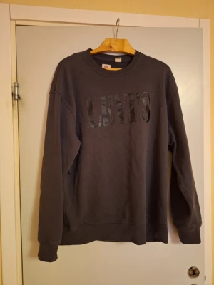Mörkgrå sweatshirt från Levi's - Säljer en mörkgrå sweatshirt från Levi's i relaxed fit. Tröjan har rund halsringning, långa ärmar och en stor svart Levi's-logga framtill. Perfekt för dig som gillar en enkel och stilren look. Materialet är mjukt och skönt, troligtvis bomull.