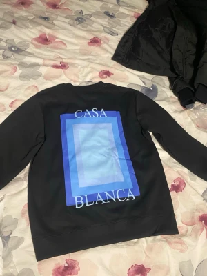 Svart sweatshirt från Casablanca - Säljer en svart sweatshirt från Casablanca med stort blått grafiskt tryck och texten 'Casa Blanca' på ryggen samt mindre tryck framtill. Tröjan har rund hals och långa ärmar. Perfekt för dig som gillar stilrena men ändå unika plagg.