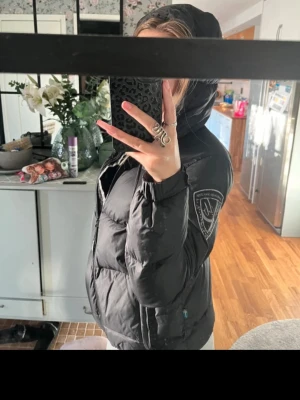 Svart pufferjacka med huva - Säljer en svart pufferjacka med huva och dragkedja framtill. Jackan har en patch på ärmen. Den är vadderad och har en normal passform, perfekt för kyliga dagar.