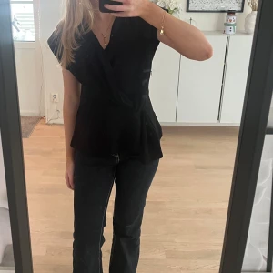 Svart omlottopp med knyt - Säljer en svart omlottopp med knyt i midjan och v-ringad hals. Toppen har korta ärmar och en lätt figurnära passform. Perfekt att styla med jeans eller kjol.