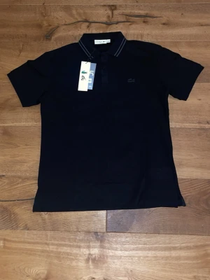 Svart pikétröja från Lacoste XL men passar L/M - Klassisk svart pikétröja från Lacoste i storlek XL. Tröjan har korta ärmar, krage med tunna vita ränder och knappar framtill. Tillverkad i bomull och har den ikoniska krokodilloggan broderad på bröstet. Perfekt för en stilren och sportig look.