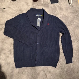 Mörkblå cardigan  RLP - Mysig mörkblå kofta från Polo Ralph Lauren med ribbstickad struktur och klassisk röd logga broderad på bröstet. Koftan har knäppning framtill, två fickor och en bred krage. Perfekt för lager-på-lager under kyliga dagar.