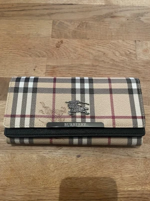 Rutig plånbok från Burberry - Säljer en klassisk plånbok från Burberry med det ikoniska rutiga mönstret i beige, svart, vitt och rött. Plånboken har flera kortfack, fönster för ID och detaljer i metall med Burberrys logga. Utsidan är i tåligt material och insidan är svart.