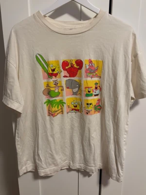 Vit Spongebob t-shirt med tryck - Vit t-shirt med färgglatt Spongebob-tryck framtill som visar flera karaktärer i olika rutor. Klassisk passform och rund hals. Står inte storlek men skulle säga S/M