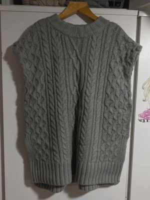 Grå stickad väst med flätmönster - Säljer en grå stickad väst med grovt flätmönster och bred ribbad kant nedtill och vid halsen. Västen är ärmlös och har en loose passform, perfekt att slänga över en skjorta eller långärmad tröja. Det finns en brun penn fläck som kan ses på bilden.