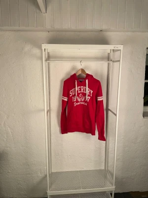 Röd Superdry hoodie med tryck - Snygg röd hoodie från Superdry med vit tryckt logga och text framtill. Tröjan har huva med vita snören, ribbade muddar och ficka på magen. Detaljer med vita ränder på ärmarna ger en sportig vibe. Mjuk insida i bomullsmix, perfekt för chill eller häng.