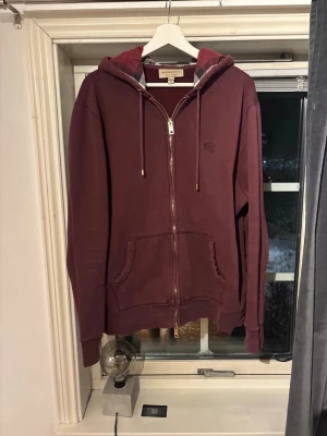 Burberry Clarendon zip hoodie - Burberry herrtröja i storlek XXL, men passar mer som L/XL. Tröjan har huva och dragkedja framtill samt en innerficka med rutigt mönster. Materialet är mjukt och lämpar sig för vardagligt bruk. Tröjan är varsamt använd men det finns täcken på slitage, såsom på metall detaljerna, samt ett ypperligt litet hål vid sidan om fickan.   Hör gärna av er om ni har frågor! Pris kan diskuteras vid snabb affär!