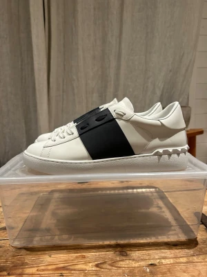 Valentino Garavani Open sneakers vit/svart - Säljer ett par feta Valentino Garavani Open sneakers i vitt skinn med bred svart läderdetalj över snörningen. Klassisk rund tå, vita snören och coola nitar på hälen. Snygg och clean design som sticker ut utan att bli för mycket.