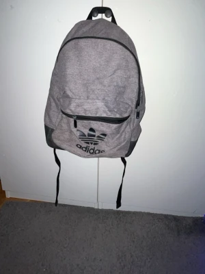 Grå ryggsäck från Adidas - Säljer en stilren grå ryggsäck från Adidas med det klassiska Trefoil-loggan i svart framtill. Väskan har ett stort huvudfack, en framficka med dragkedja och justerbara axelband. Materialet är slitstark polyester och designen är enkel men sportig. Denna Adidas väska är i nytt skick och sparsamt använd. Nypris 300 kr jag säljer för 50.