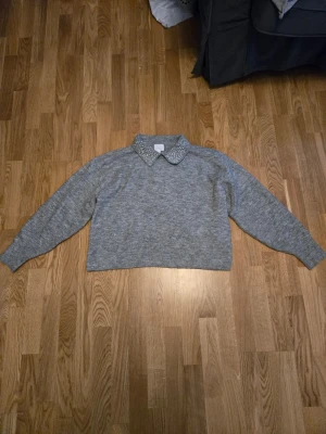 Grå stickad tröja med pärlkrage H&M - Säljer en grå stickad tröja från H&M i storlek L. Tröjan har lång ärm och en snygg krage dekorerad med pärlor som ger en extra touch. 