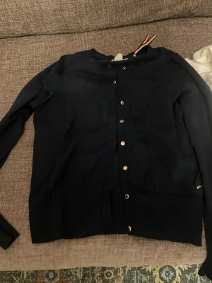 Mörkblå kofta från Lindex  - Snygg mörkblå kofta från Holly & Whyte i storlek M. Koftan har långa ärmar, rund hals och guldfärgade knappar framtill. Materialet är mjukt och stickat, perfekt för lager på lager under kyliga dagar. Använd ett fåtal gånger 