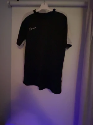 Svart Nike Dri-FIT t-shirt 158-170 - Säljer en svart Nike Dri-FIT t-shirt i storlek 158-170 med vita detaljer längs axlar och sidor. Tröjan har klassisk rund hals och är tillverkad i ett lätt, snabbtorkande material som passar perfekt för träning eller sport. 