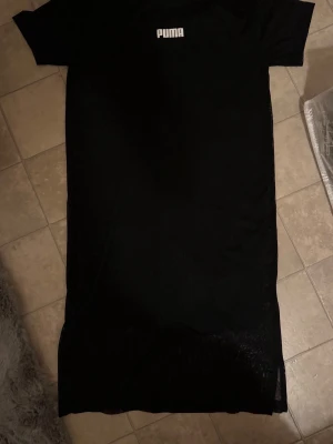 Svart klänning från Puma - Svart klänning från Puma med klassisk rund halsringning och korta ärmar. Klänningen har en enkel och sportig look med Puma-logga framtill. Tillverkad i ett lätt och bekvämt material som passar perfekt för en avslappnad stil.