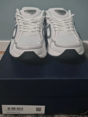 Vita Dior B30 sneakers - Säljer ett par riktigt snygga Dior B30 sneakers i vitt med gråa detaljer och stor CD-logga på sidan. Skorna har meshpaneler för extra andningsförmåga och en chunky sula som ger grym streetstyle-vibe. Klassisk snörning och exklusiv känsla, perfekta för dig som gillar lyxiga sneakers.