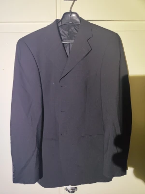 Svart kavaj från Batistini - Säljer en klassisk svart kavaj från Batistini. Kavajen har enkelknäppning, snygg krage och raka fickor framtill och knappar. Materialet är slätt och känns stilrent, perfekt till skjorta eller t-shirt. Långärmad modell med knappar vid ärmslut.