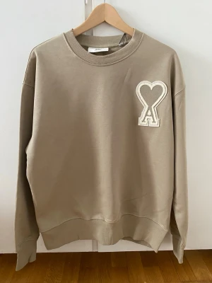 Ami Paris Sweatshirt – Beige - Storlek: M Skick: Ny & oanvänd  Beige Ami Paris sweatshirt med broderad Ami de Cœur-logga på bröstet. Tillverkad i mjuk bomullsblandning med ribbad krage, muddar och nederkant. Rak och modern passform – lätt att styla till allt. Pris kan diskuteras om du är intresserad!