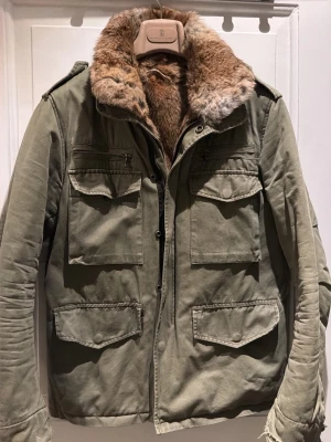 Olivgrön fieldjacka med äkta kaninpälspäls - Säljer en olivgrön fieldjacka med stor, mjuk äkta kaninpäls på krage och insida. Jackan har fyra stora fickor framtill, axelklaffar och dragkedja. Perfekt för kalla dagar och ger en riktigt snygg och avslappnad look.