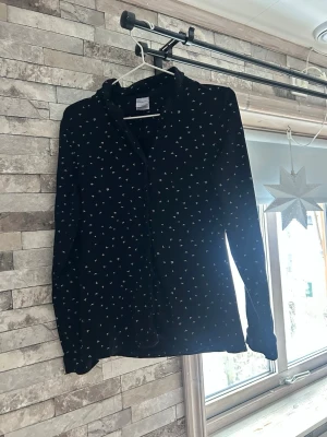 Svart skjorta med små vita fåglar - Snygg svart skjorta  med små vita broderade fåglar över hela tyget. Skjortan har lång ärm, klassisk krage och knäppning framtill. Tillverkad i mjuk bomull som känns skön mot huden. Perfekt för dig som gillar detaljer och stilren design. Sov plagg 